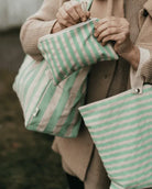 Eine Person hält die Pocket Reissverschlusstasche von OMOM und eine Tragetasche, beide von Main Design und aus Bio-Baumwolle, während sie einen beigen Mantel und Pullover trägt.