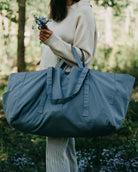 Eine Person in einem cremefarbenen Pullover und einer gestreiften Hose steht draußen im Grünen, hält kleine Blumen und trägt die Stofftasche gross von OMOM by Main Design.