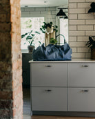 Eine blaue Stofftasche gross von OMOM by Main Design steht auf einem hellgrauen Küchentisch in einer modernen Küche mit weißen Subway-Fliesen, Topfpflanzen und natürlichem Licht, das durch das Fenster fällt.