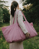 Eine Frau mit Pferdeschwanz und weißem Pullover steht im Freien auf einer Wiese und hält die "Stofftasche gross von OMOM" von Main Design über ihre Schulter. Im Hintergrund sind Sonnenlicht und Bäume zu sehen.