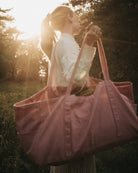 Eine Frau mit Pferdeschwanz und weißem Pullover steht im Freien im Sonnenlicht und trägt die Stofftasche gross von OMOM von Main Design über der Schulter. Im Hintergrund sind Bäume und Gras zu sehen.