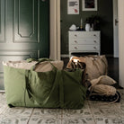 Ein grüner Seesack, die Stofftasche gross von OMOM by Main Design, und eine Kordeltasche liegen auf einem gefliesten Boden neben einer offenen Tür, mit einer weißen Kommode und einer grünen Wand im Hintergrund.