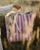 Eine Person, die einen hellen Pullover trägt, steht in einem sonnenbeschienenen Feld und hält eine Stofftasche von OMOM by Main Design mit einer gewebten beigen Decke darin.