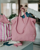 Eine Person hält eine rosa Stofftasche von OMOM von Main Design, die mit Blattgrün gefüllt ist, während eine andere rosa gestreifte Stofftasche von OMOM und Stoffbeutel auf einem Küchentisch im Hintergrund liegen.