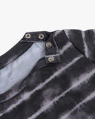 Nahaufnahme eines schwarz-grauen Batik-UV-Shirts von Aina für Kinder aus nachhaltigem Stoff mit einzigartigem Ausschnittdesign mit Druckknöpfen von Main Design. Die Druckknöpfe sind offen dargestellt und geben den Blick auf die Innenseite des Shirts frei. Der Stoff hat eine weiche Textur und das Batikmuster ist horizontal.