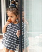 Ein kleines Kind mit einem dunklen Haargummi steht neben einer Glastür und nuckelt an einem Schnuller. Sie trägt ein schwarz-weiß gestreiftes Aina Kinder UV-Shirt von Main Design mit UPF 50+ Sonnenschutz und blickt mit nachdenklichem Gesichtsausdruck nach draußen. Ihr Spiegelbild ist im Glas neben ihr zu sehen.