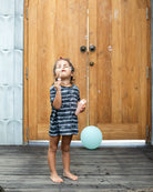 Ein kleines Kind in einem Main Design Aina Kinder UV-Shirt steht auf einem Holzdeck und macht Seifenblasen vor einer großen Holztür. Hinter ihnen ist an der Türklinke ein blauer Ballon befestigt. Das Kind scheint entzückt zu sein, während es den Seifenblasen beim Schweben zusieht.