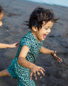 Ein kleines Kind mit unordentlichem Haar und sandigen Händen läuft verspielt am Strand in einem grün gemusterten Aina Kinder UV Shirt von Main Design, das schnelltrocknend ist und UPF 50+ Schutz bietet. Ein weiterer Kinderarm erscheint in der Nähe.