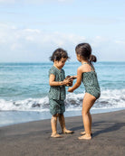 Zwei Kinder in passenden grünen Aina Kinder UV Shirts von Main Design (UPF 50+, schnelltrocknend) spielen und lächeln an einem Sandstrand mit Wellen und blauem Himmel im Hintergrund, ihre Beine sind mit Sand bedeckt.