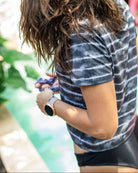 Frau mit langem, welligem Haar, trägt ein Main Design Lush UV Surf Shirt und eine Smartwatch und steht im Freien vor einem verschwommenen grünen und rosa Hintergrund. Sie hält einen Gegenstand in den Händen, möglicherweise eine Schere oder ein Gartengerät.