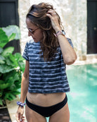 Eine Frau mit Sonnenbrille, einem Lush UV Surf Shirt von Main Design und einem schwarzen Bikinihöschen, steht an einem Pool. Sie hat eine Hand in ihrem Haar und im Hintergrund sind üppige grüne Pflanzen und Steinmauern zu sehen.