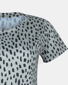 Nahaufnahme des Lush UV Surf Shirts von Main Design in Hellgrau mit unregelmäßigem schwarzem Tupfenmuster und Rundhalsausschnitt. Dieses kurzärmelige, schnell trocknende Oberteil ist von der Schulter bis zur Mitte des Oberkörpers vor einem einfarbig weißen Hintergrund abgebildet.