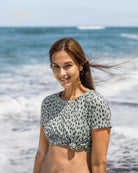 Eine Frau mit langen braunen Haaren und goldenen Ohrringen steht an einem Strand und lächelt in die Kamera. Sie trägt das Main Design Lush UV Surf Shirt - ein schnelltrocknendes, UPF 50+ beschnittenes Oberteil mit einem Frontknoten. Wellen und blauer Ozean sind hinter ihr.