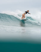 Eine Frau, die das Main Design Maria Langarm Lycra-Oberteil mit UPF 50+ und eine Bikinihose trägt, surft auf einer Welle und balanciert mit gebeugten Knien und ausgestreckten Armen über das klare blaugrüne Wasser.