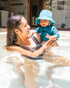 Eine Frau in einem schwarzen Badeanzug lächelt, während sie ein Baby in einem blaugrünen Sonnenhut und der Main Design Marlon Kinder Lycra-Badebekleidung mit UPF 50+ Schutz hält, während sie einen sonnigen Tag in einem flachen Freibad genießen.
