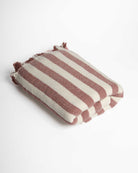 Das Strandtuch Melody von Main Design ist ein gefaltetes, rechteckiges Hamam-Handtuch aus recycelter Baumwolle mit breiten kastanienbraunen und cremefarbenen Streifen mit ausgefransten Rändern auf weißem Hintergrund.