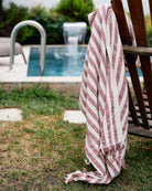 Das rosa-weiß gestreifte Strandtuch Melody von Main Design aus recycelter Baumwolle hängt über einem Holzstuhl neben einem Swimmingpool, mit grünem Gras und einem Wasserspiel im Hintergrund.