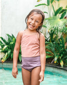 Ein junges Mädchen mit nassen Haaren lächelt strahlend an einem Pool in dem Lena Mädchen Badeanzug von Main Design. Üppige grüne Pflanzen und tropische Blätter im Hintergrund schaffen eine fröhliche, sommerliche Atmosphäre.