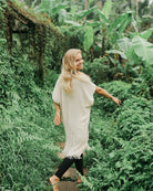 Eine Frau mit langen blonden Haaren, bekleidet mit einem hellbeigen Kimono Saturn von Main Design und schwarzen Leggings, geht barfuß einen üppig grünen, tropischen Weg entlang und lächelt über ihre Schulter zurück.