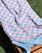 Ein „Strandtuch Pixel“ von Main Design mit rosa-blauen Karos liegt über einem Liegestuhl aus Holz auf grünem Gras, während das Sonnenlicht sanfte Schatten auf den Stoff aus recycelter Baumwolle wirft.