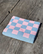 Das Strandtuch „Pixel“ von Main Design aus recycelter Baumwolle mit pink-blauem Schachbrettmuster liegt auf wettergegerbtem Holz im hellen Sonnenlicht – modernes Design für entspannte Tage.