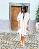 Eine barfüßige Frau mit nassen Haaren steht auf Kopfsteinpflaster vor einer Holztür und trägt einen schwarzen Bikini mit dem hell gestreiften Kimono Naya von Main Design als Fransenüberwurf - das perfekte Sommer-Outfit.