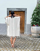 Eine barfuß gehende Person mit nassen Haaren trägt den weißen Kimono Naya von Main Design. Sie steht auf Pflastersteinen in der Nähe einer Holztür und eines großen Pflanzkübels mit grünen Pflanzen und bunten Blumen, die Hände zum Kopf erhoben.