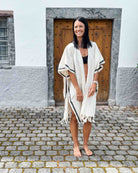 Eine Frau mit langen dunklen Haaren trägt den Kimono Naya von Main Design über einem schwarzen Bikini und steht barfuß auf einem Kopfsteinpflasterweg vor einer Holztür in einem weißen Gebäude. Sie lächelt und blickt zur Seite.