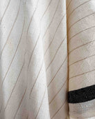 Eine Nahaufnahme des Kimono Naya von Main Design zeigt cremefarbenen Stoff mit dünnen diagonalen beigen Streifen und einem dicken schwarzen Streifen am Rand. Das strukturierte Gewebe aus OEKO-TEX-zertifizierter Baumwolle ist ideal für einen stilvollen Strandkimono.
