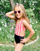 Ein lächelndes Mädchen mit rosa Sonnenbrille und dem Milly Mädchen Badeanzug von Main Design mit UPF50+ steht mit einer Hand in der Hüfte in einem Garten mit grünen Pflanzen und weißen Blumen.