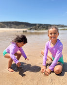 Zwei kleine Kinder in einem passenden Main Design Laila Mädchen Badeanzug mit UPF 50+ spielen an einem Sandstrand in der Nähe des seichten Wassers. Im Hintergrund sind grasbewachsene Hügel und Felsen zu sehen, und der Himmel ist blau.