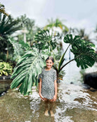 Ein junges Mädchen im Main Design Aina Kinder UV Shirt steht barfuß im flachen Wasser inmitten tropischer Blätter und lächelt in die Kamera. Das Shirt mit UPF 50+ ist ideal für ihr sonnenreiches Outdoor-Abenteuer.