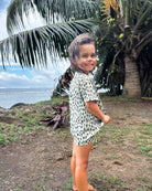 Ein kleines Kind mit langen Haaren lächelt barfuß im Gras in der Nähe des Ufers und trägt ein Main Design Aina Kinder UV Shirt. Große Palmen und das Meer sind im Hintergrund unter einem teilweise bewölkten Himmel zu sehen.