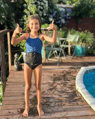 Ein Mädchen trägt den Lena Mädchen Badeanzug von Main Design. Sie steht lächelnd auf einem Holzdeck am Pool, zeigt zwei Daumen hoch. Im Hintergrund sind Outdoor-Möbel und grüne Pflanzen bei sonnigem Wetter zu sehen.