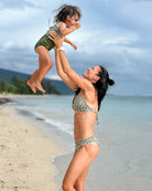 Eine Frau hebt am Sandstrand ein lachendes Kind hoch, das den Lena Mädchen Badeanzug von Main Design trägt. Im Hintergrund rahmen Palmen und ein bewölkter Himmel die fröhliche Szene am Meer.