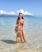 Eine Frau und ein junges Mädchen, die beide das Drift Surf Bikini Oberteil von Main Design mit passendem Leopardenmuster tragen, lächeln zusammen, während sie im klaren, seichten Ozeanwasser stehen, mit Bergen und blauem Himmel im Hintergrund. all-groups