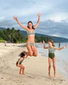 Eine Frau in einem nachhaltigen Badeanzug springt freudig an einem Sandstrand mit zwei Mädchen, die Lena Mädchen Badeanzug von Main Design tragen. Im bewölkten Hintergrund sind Palmen und Berge zu sehen.