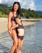 Eine Frau und ein junges Mädchen, beide in passenden dunkelvioletten Nalu Mädchen Bikinis von Main Design (UPF 50+), stehen zusammen an einem sonnigen Sandstrand mit seichtem Wasser und üppigem Grün im Hintergrund.