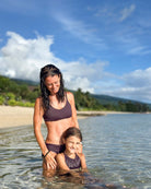 Eine Frau in einem schwarzen Sassy Bikini-Top von Main Design, hergestellt aus recycelten Materialien, steht lächelnd mit den Händen auf den Schultern eines jungen Mädchens im klaren Strandwasser. Beide tragen umweltfreundliche Badeanzüge inmitten von üppigem Grün und blauem Himmel.
