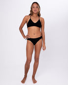 Eine Frau steht barfuß vor einem weißen Hintergrund und trägt die Serene Surf Bikini Hose von Main Design, die aus recycelten Materialien hergestellt wurde. Eine Hand ruht auf ihrer Hüfte, während sie mit neutralem Blick in die Kamera schaut.