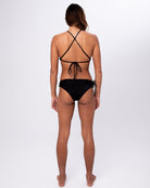 Eine Frau steht barfuß, den Blick abgewandt, und trägt einen schwarzen Bikini mit überkreuzten Trägern und Serene Surf Bikini Hose von Main Design, hergestellt aus recycelten Materialien, vor einem schlichten weißen Hintergrund.