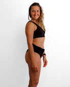 Eine Frau mit langem, gewelltem Haar steht seitlich und lächelt. Sie trägt die schwarze Alice Bikini Hose von Main Design aus recycelten Materialien vor einem schlichten weißen Hintergrund.
