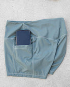Ein Paar „Aura Bike Shorts“ von Main Design in Hellblau mit mittelhohem Bund und verdeckten Seitentaschen – abgebildet auf einem hellgrauen Hintergrund, wobei ein Handy teilweise in eine der Taschen gesteckt ist.