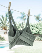 Ein grünes „Essence Active“-Top von Main Design aus recycelten Materialien hängt mit Wäscheklammern an einer Wäscheleine. Im Hintergrund sind Palmen, Büsche und ein Swimmingpool in einer sonnigen Außenkulisse zu sehen.