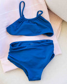 Der „Nalu Mädchen Bikini“ von Main Design, ein blauer Zweiteiler mit einem kurz geschnittenen Oberteil und einer hoch taillierten Hose, der aus recycelten Materialien hergestellt ist und Sonnenschutz der Klasse UPF 50+ bietet, liegt auf gefalteten, hellen Handtüchern.