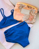 Ein leuchtend blauer „Nalu Mädchen“-Bikini von Main Design, der aus recycelten Materialien gefertigt ist und Sonnenschutz der Klasse UPF 50+ bietet, liegt auf einem hellrosa Handtuch neben einer Tasche mit Reißverschluss, die mit einem tropischen Blumenmuster und einem „ALOHA“-Etikett verziert ist – perfekt für unbeschwerte Tage in der Sonne.