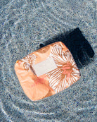 Ein kleines Täschchen aus der Aloha-Kollektion mit einem pfirsichfarbenen Blumenmuster und einem strahlend weißen „ALOHA“-Anhänger schwimmt auf kristallklarem, plätscherndem Wasser, dessen sanfter Schatten sich darunter abzeichnet – Ihre perfekte wasserdichte Tasche und modische Bikini-Tasche für jedes Abenteuer am Pool.