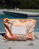 Ein kleines Täschchen aus der Aloha-Kollektion mit einem Blumenmuster in Pfirsich- und Elfenbeintönen liegt am Rand eines Pools, umgeben von verschwommenem Grün und glitzerndem Wasser an einem sonnigen Tag. Dieses stilvolle, wasserabweisende Täschchen aus der Aloha-Kollektion eignet sich perfekt als Bikini-Tasche, um Ihre wichtigsten Utensilien am Wasser trocken und ordentlich aufzubewahren.