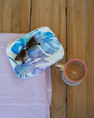 Auf einem Holztisch steht eine rosa Kaffeetasse neben einem zusammengefalteten rosa Tuch, einer „Aloha Collection Aloha Small Pouch“ – die sich mit ihrem blau-weißen Blattmuster ideal als Bikini-Tasche oder wasserdichte Tasche eignet – und einer darauf liegenden Sonnenbrille.