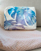 Ein weißes Kissen mit einem blau-lila Blattmotiv im Aquarellstil liegt auf einem grauen, strukturierten Kissen und einer rosa Decke mit Chevron-Muster, dazu gehört die „Aloha Collection Aloha Small Pouch“ – die ideale wasserdichte Tasche, um Ihre wichtigsten Utensilien stilvoll zu verstauen.